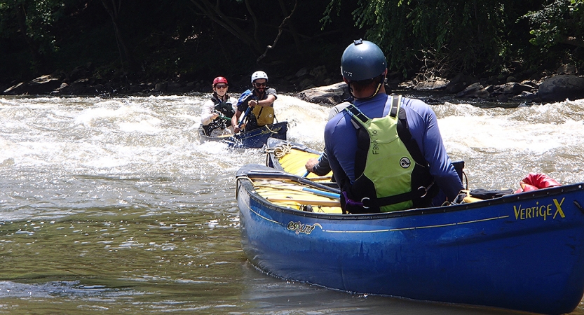 Two students paddle a canoe through whitewater, while another person who is likely an instructor watches them from another canoe.